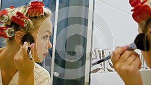 A beautiful young woman looks in the mirror and applies powder to her face. Cute blonde in curlers on her head getting