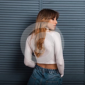 Beautiful young woman with luxurious red long hair in a stylish white shirt in jeans stands near a vintage blue wooden wall
