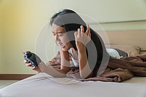 Beautiful young woman lying on a bed holding an alarm clock in her hand with exasperation