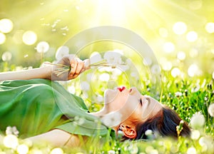 Beautiful young woman lying on the field in green grass and blowing dandelion flowers