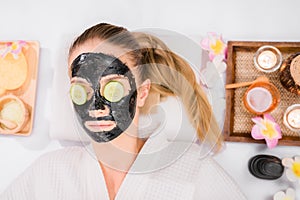 Beautiful young woman lying on spa bed facial mask by black cosmetic and use cucumber slices on eyes in spa salon