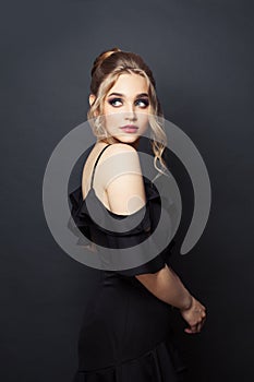 Beautiful young woman with makeup and blonde hairdo wearing black dress on black background