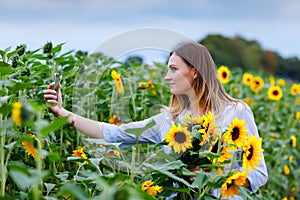 Beautiful young woman making selfie with smartphone on sunflower field with bouquet flowers. Happy girl on summer sunset