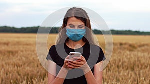 Beautiful young woman in a medical mask to protect against the virus, standing in a field and looking at the phone.