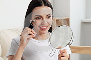 Beautiful young woman with mirror using eyelash curler indoors