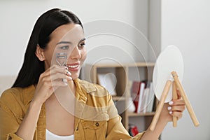 Beautiful young woman with mirror using eyelash curler indoors