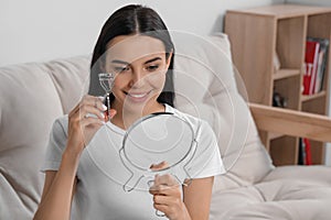 Beautiful young woman with mirror using eyelash curler indoors