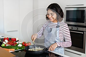 Beautiful young woman mixing rice with vegetables in frying pan