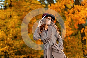 Beautiful young woman model with a cute smile in a stylish coat in a fashionable hat in trendy glasses stands in the park and