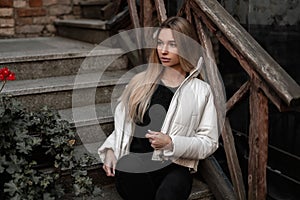 Beautiful young woman model in fashionable black-white outerwear relaxes on a stone old staircase with a wooden railing.