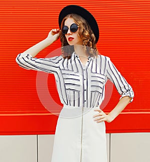 Beautiful young woman model wearing white striped shirt, black round hat posing on city street over red wall