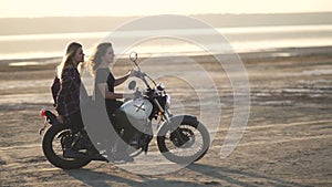 Beautiful young woman motorcyclist with his girlfriend riding a motorcycle in a desert on sunset or sunrise. Girlfriends on a bike