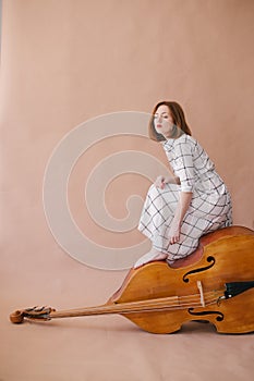 Beautiful young woman musician sitting on a vintage double bass on a beige background