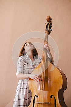 Beautiful young woman musician sitting on a vintage double bass on a beige background