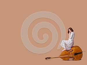 Beautiful young woman musician sitting on a vintage double bass on a beige background