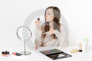 Beautiful young woman painting nails with red nail polish, sitting at table applying makeup with set facial decorative