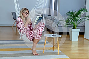 Beautiful young woman in pajamas reading a book in hammock chair.