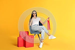 Beautiful young woman with paper shopping bags in armchair on yellow background