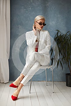 Beautiful young woman with perfect blond hair in elegant white suit and red shoes posing in studio. Fashionable blonde