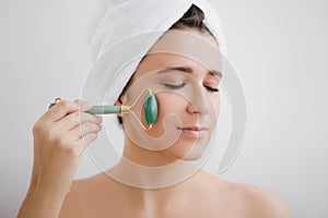 Beautiful young woman with perfect skin wearing towel on head using a jade face roller with natural quartz stones