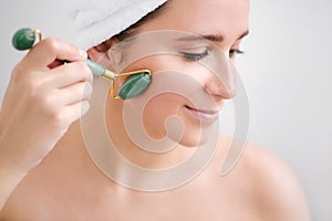 Beautiful young woman with perfect skin wearing towel on head using a jade face roller with natural quartz stones