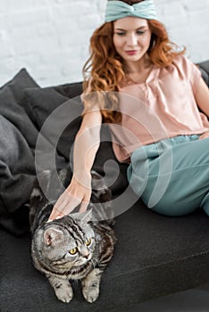 beautiful young woman petting tabby cat while sitting on couch