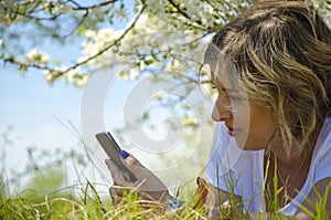 Beautiful young woman with a phone, lying on the field, green grass and flowers. Outdoors enjoy nature. Healthy smiling girl lying