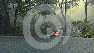 Beautiful young woman in pink bikini enjoying tropical rain falling on her in infinity pool with mystic jungle view