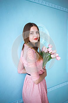 Beautiful young woman in pink dress holding in hands spring tulips flowers bouquet on blue wall background