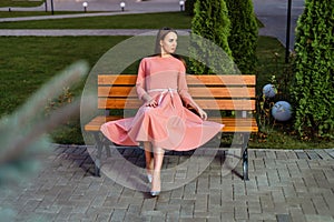 Beautiful young woman in pink dress is sitting on a wooden bench.
