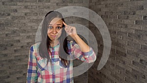 A beautiful young woman in a plaid shirt twists at her temple on a gray brick background.