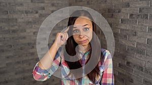 A beautiful young woman in a plaid shirt twists at her temple on a gray brick background.