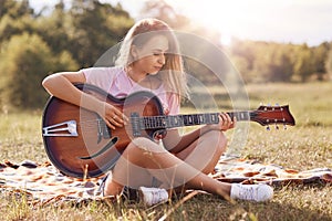 Beautiful young woman playing gitar in meadow, has stright blonde hair, looks concentrated on musical insrtument, wearing casual