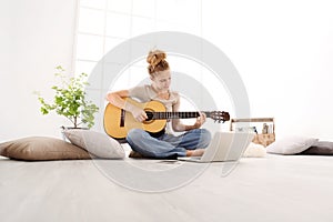 Beautiful young woman playing guitar with computer, learn to play with an online course, sitting on the floor in casual clothes