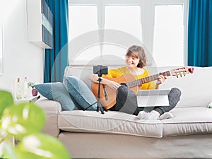 Beautiful young woman playing guitar with laptop at home. online course to learn play guitar. blogger girl sitting on