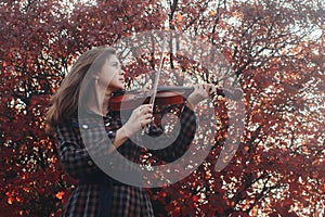 Beautiful young woman playing violin on a background of red foliage, romantic girl in dress playing a musical instrument in nature