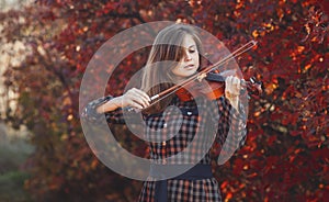 Beautiful young woman playing violin on a background of red foliage, romantic girl in dress playing a musical instrument in nature