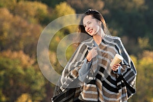Beautiful young woman in a poncho with coffee in hand on nature background. Brunette girl laughing