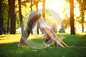 Beautiful young woman practices yoga asana downward facing dog in the park at sunset