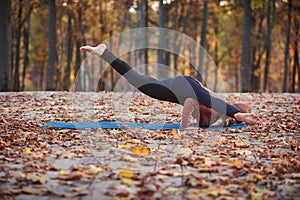 Beautiful young woman practices yoga asana Eka Pada Kaundiniasana 2 on the wooden deck in the autumn park