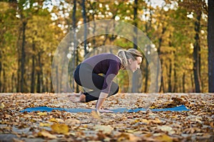 Beautiful young woman practices yoga asana Lolasana Pendant pose on the wooden deck in the autumn park.