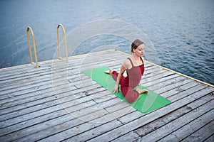 Beautiful young woman practices yoga asana One Legged King Pigeon Pose - Eka Pada Rajakapotasana on the wooden deck near the lake