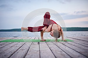 Beautiful young woman practices yoga asana Parivritta Eka Pada Danda Kaundiniasana on the wooden deck near the lake