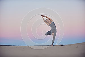 Beautiful young woman practices yoga asana Parshva Vrikshasana - Tree pose in the desert at sunset