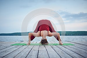 Beautiful young woman practices yoga asana Prasarita Padottanasana on the wooden deck near the lake