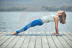 Beautiful young woman practices yoga asana Purvottanasana Upward Plank Pose on the wooden deck near the lake