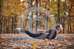 Beautiful young woman practices yoga asana Salabhasana locust pose on the wooden deck in the autumn park