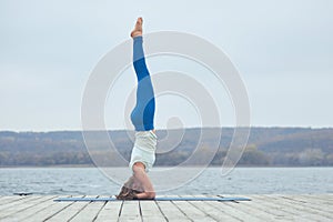 Beautiful young woman practices yoga asana Shirshasana - Headstand pose on the wooden deck near the lake