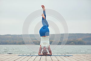 Beautiful young woman practices yoga asana Shirshasana - Headstand pose on the wooden deck near the lake