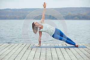 Beautiful young woman practices yoga asana Vasishthasana - side plank pose on the wooden deck near the lake